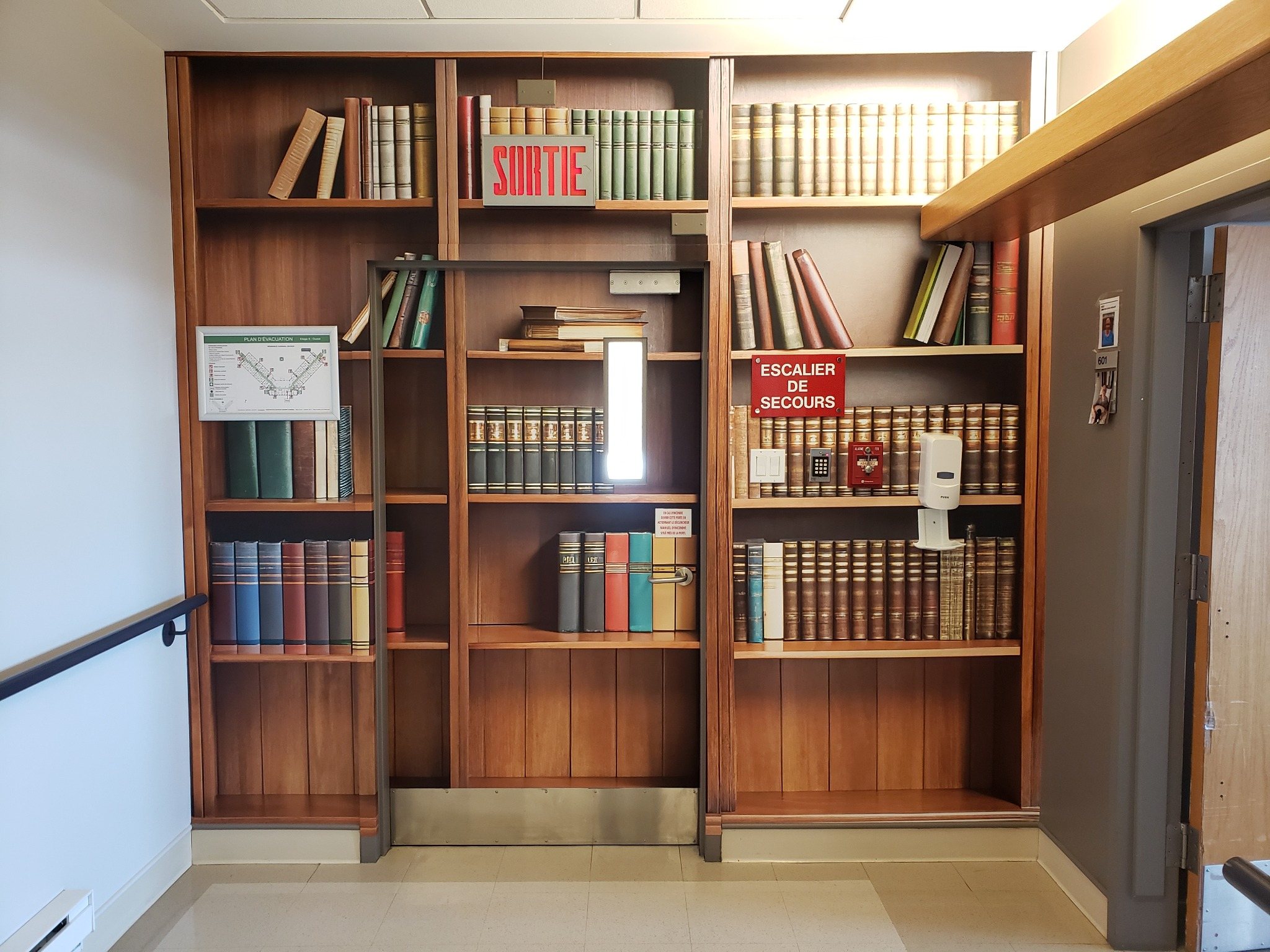 Murale en trompe-l’œil de bibliothèque couvrant un mur et une porte, avec les indications de sortie visibles.