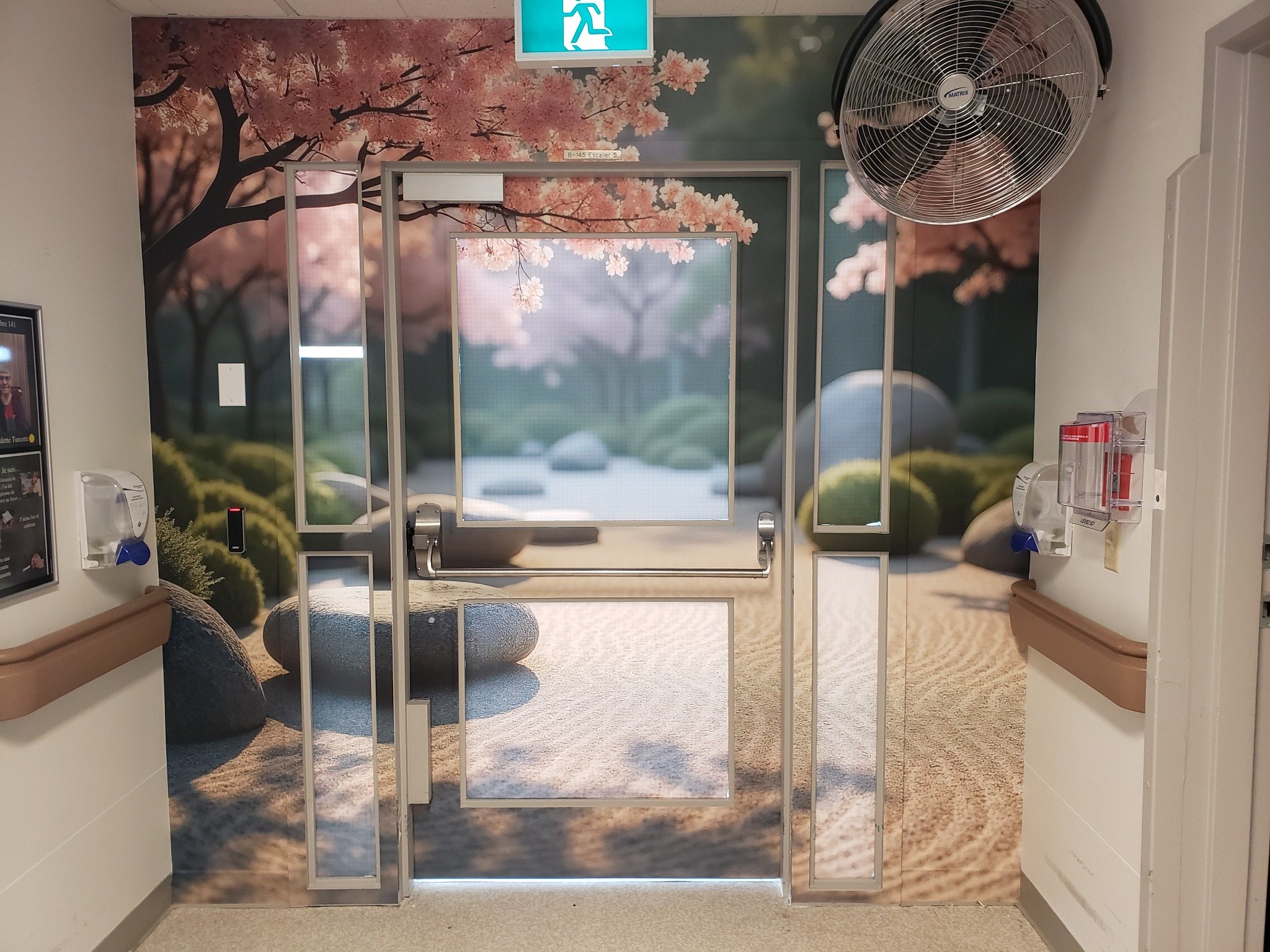 Décor mural de jardin avec cerisier en fleurs et pierres, prolongé sur une porte au bout d’un corridor avec mains courantes.